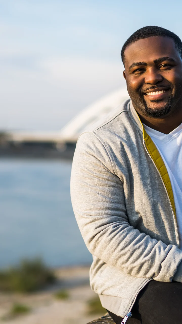 Heavier man in a gray hoodie smiling on a wall by a river with a bridge behind him