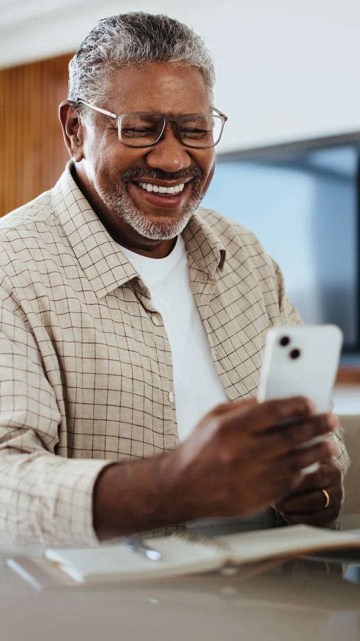 Older Black man laughing at his phone with a laptop on the desk