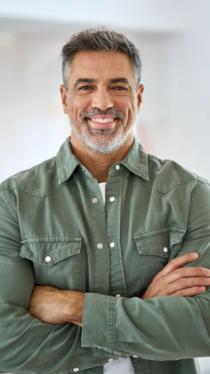 Mature gray-haired man in an olive shirt with arms crossed smiling at home