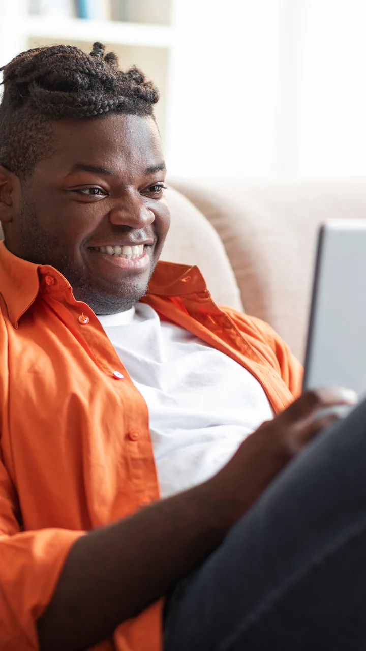 Young Black man in an orange shirt laughing while watching something on a tablet